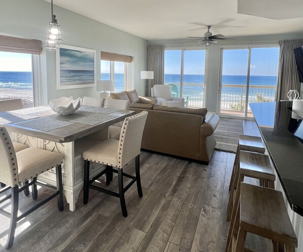 Un amplio salón con una mesa de comedor de madera, sillas tapizadas en beige, un sofá beige y un ventanal con vistas al océano.