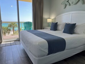 Una habitación de hotel con una cama grande, un balcón con vista a la playa y una mesita de noche con una lámpara.