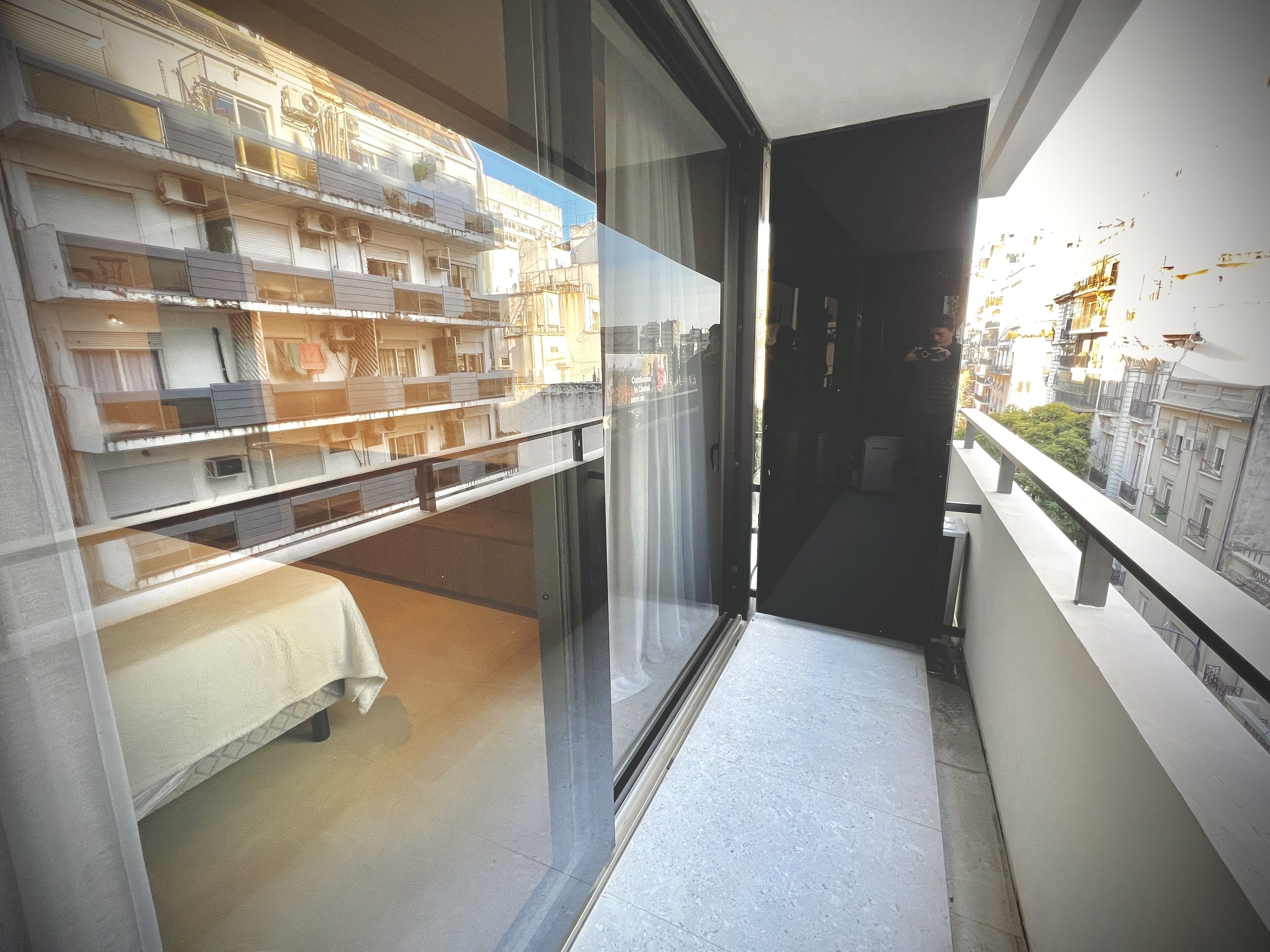 A narrow balcony with a glass door leading to a room with a bed and a small table.