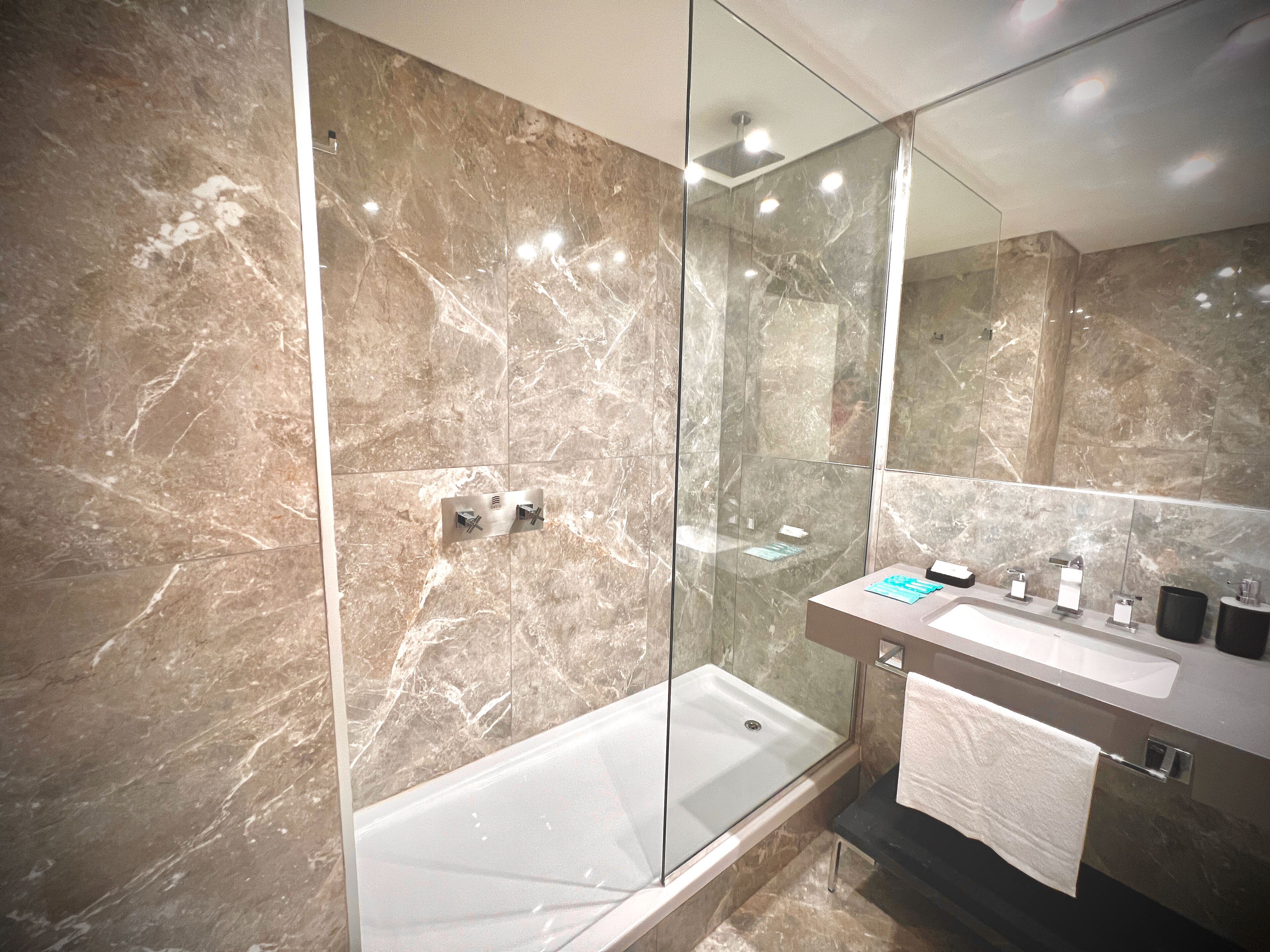 A modern bathroom with a glass-enclosed shower, a white bathtub, and a marble-tiled wall.