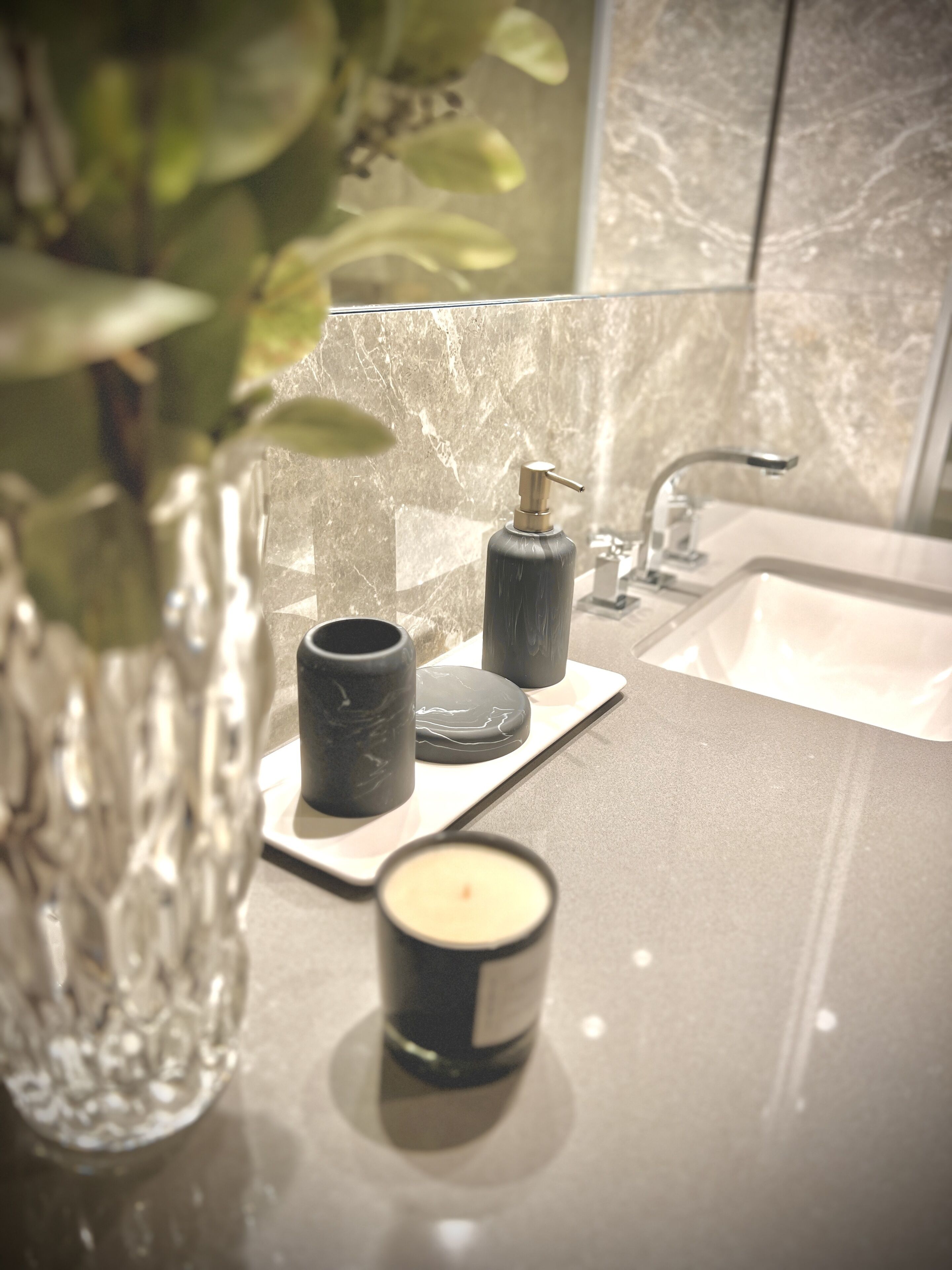 A bathroom with a marble sink, a black soap dispenser, a black candle, and a glass vase with green leaves.