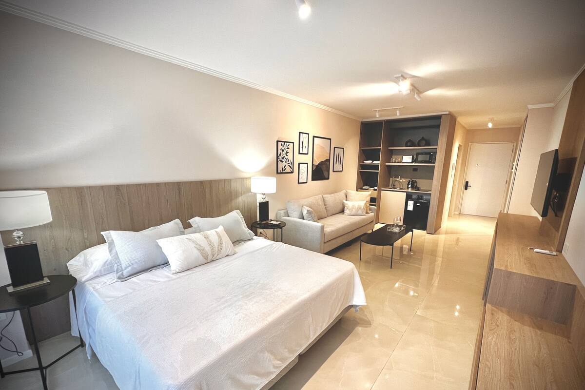 Habitació d'hotel amb llit, sofà, taula petita, escriptori, làmpada, televisió i cuina americana.