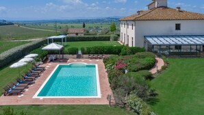 A large rectangular swimming pool with lounge chairs and umbrellas, surrounded by well-maintained lawns and hedges, with a view of distant hills and a large building with a tiled roof.