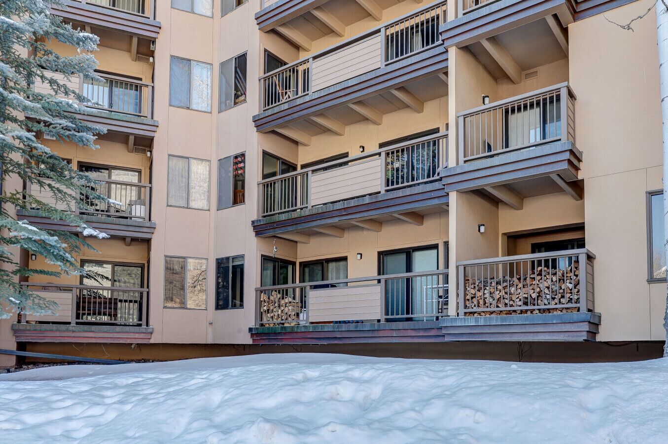 A multi-story residential building with balconies, a snow-covered ground, and a tree in the foreground.