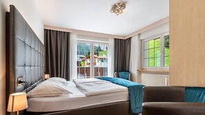 A hotel room with a bed, a chair, a lamp, a window with curtains, and a view of houses outside. - Apartment 'Mozart' with Mountain View, Balcony and Wi-Fi (Selva in Val Gardena)