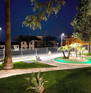 A nighttime view of a landscaped area with a pool, palm trees, and a white fence.