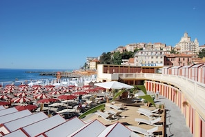 A beachside hotel with multiple sun loungers and umbrellas, a pool area, and a view of the sea and nearby buildings.
