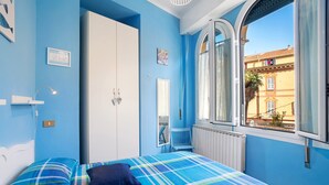 Une chambre à coucher comprenant un lit, une armoire, une chaise et une fenêtre donnant sur des immeubles.