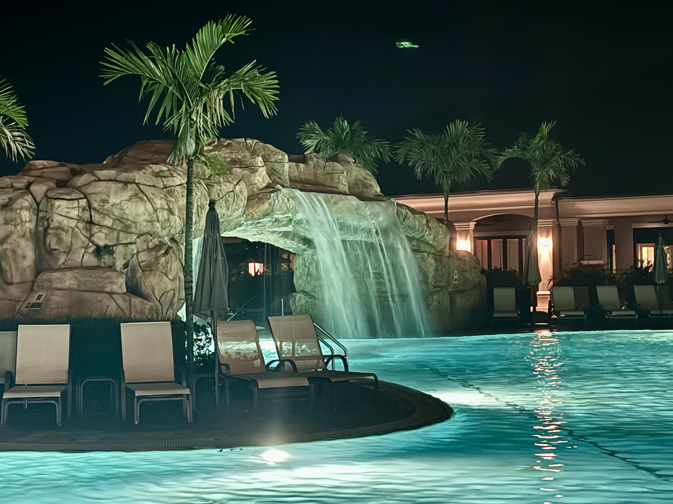 Una piscina con cascada, sillas de descanso, palmeras y un edificio al fondo.