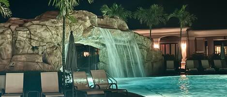Una piscina con cascada, sillas de descanso, palmeras y un edificio al fondo.