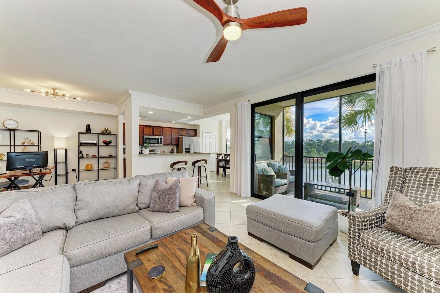 A modern living room with a sofa, ottoman, and coffee table, a kitchen in the background, and a balcony with a view.