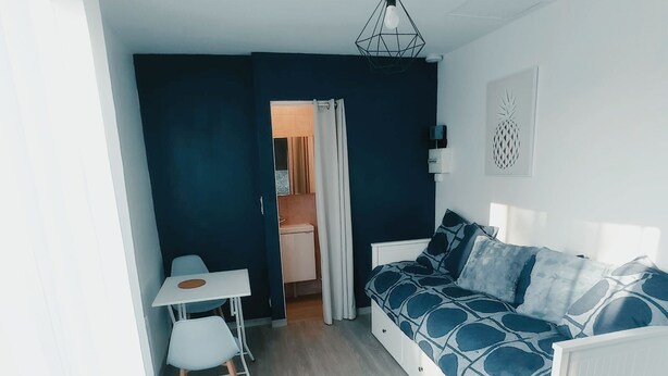 A modern, compact living space with a white sofa, a small dining table, and a kitchenette in the background.