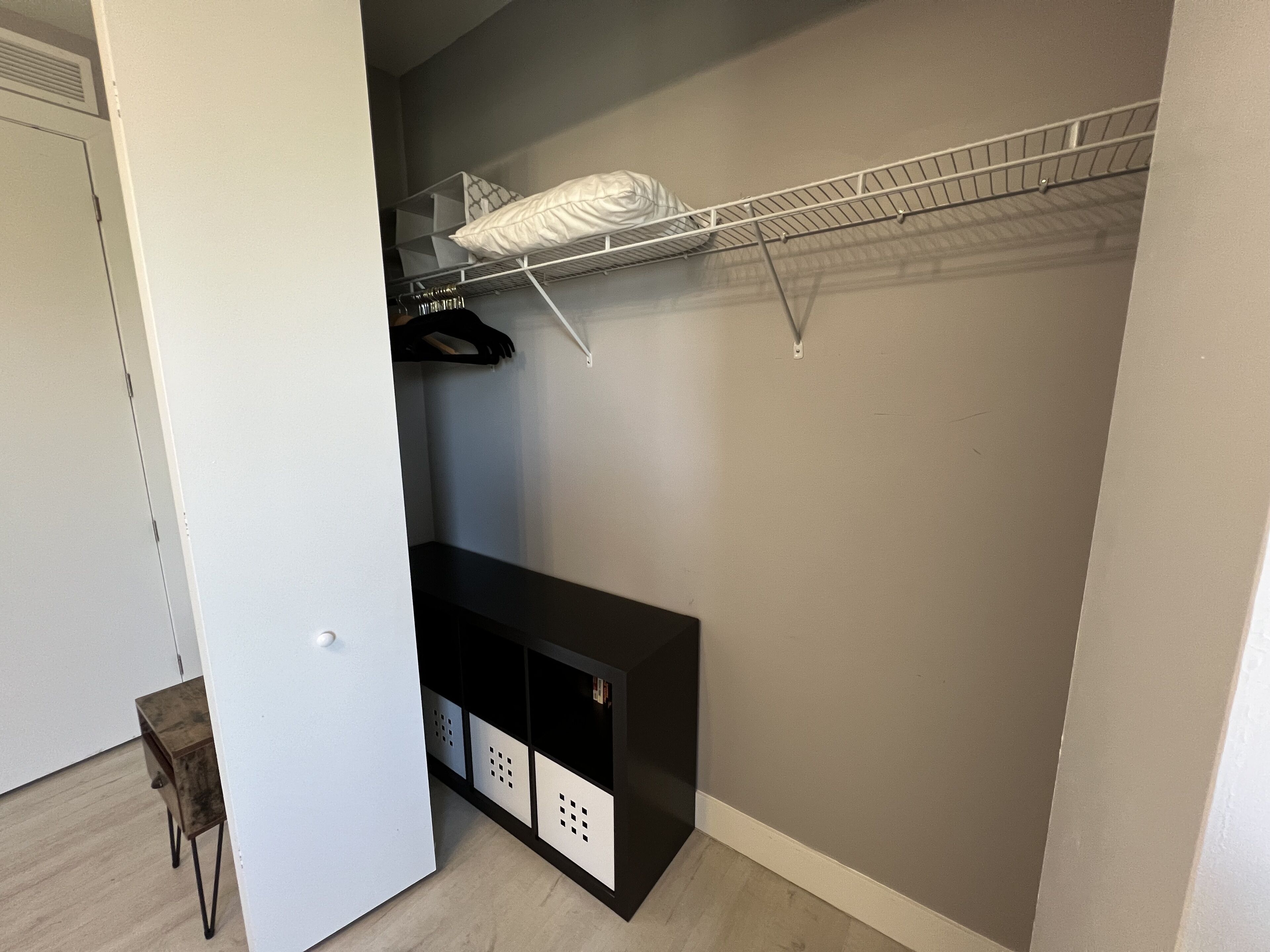 Un vestidor con un estante metálico, un banco negro para guardar objetos y una puerta blanca.