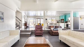 A spacious living area with a central coffee table, two sofas, and a reclining chair. A kitchen with teal cabinets and modern appliances is visible in the background. - Yosemite Lakehouse Sleeps 20 + Hot Tub + Game Room + BBQ (Oakhurst)