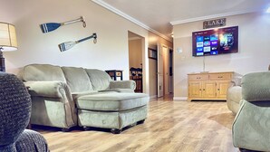 A living room with a sectional sofa, wooden flooring, a flat-screen TV mounted on the wall, and a sign that reads "LAKE". - Hidden Bliss-Large 4 bedroom home at Mt Storm Lake (Mount Storm)