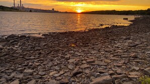 A rocky shoreline with a sunset over a body of water, industrial structures in the background, and a clear sky. - Hidden Bliss-Large 4 bedroom home at Mt Storm Lake (Mount Storm)