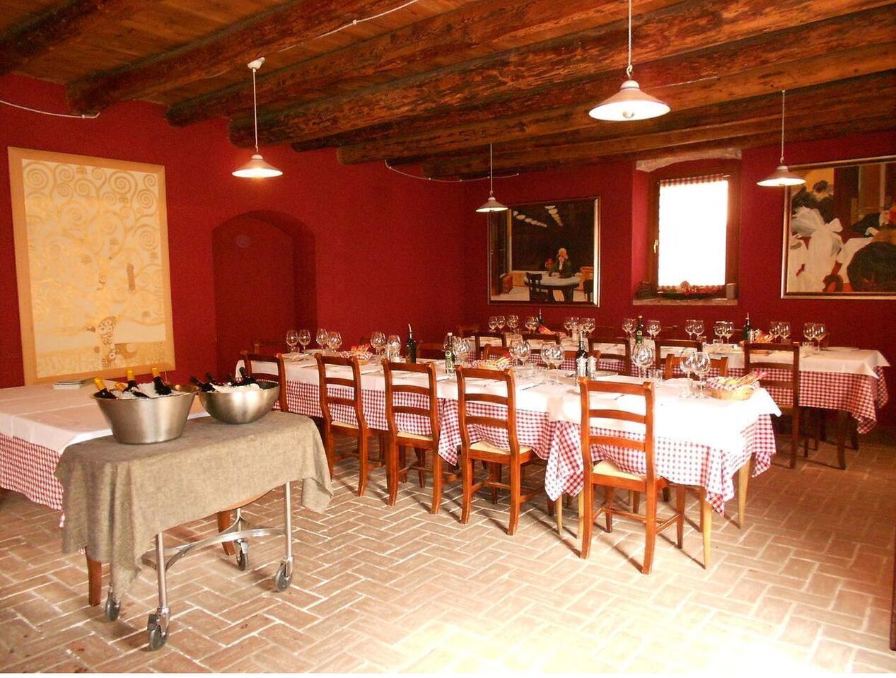 Uma sala de jantar com paredes vermelhas, vigas de madeira no teto e toalhas de mesa xadrez. Há baldes de metal com garrafas sobre uma mesa, e o chão é ladrilhado.