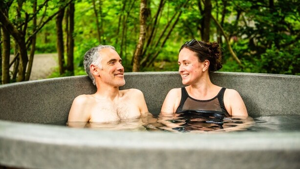 Deux personnes dans un jacuzzi extérieur, entourées d’arbres.