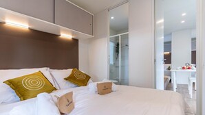 A modern hotel room with a large bed, a glass partition, and a dining area visible through the partition.