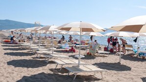A beach with rows of sun umbrellas and lounge chairs, people sunbathing and swimming, and a clear view of the sea and mountains in the background.