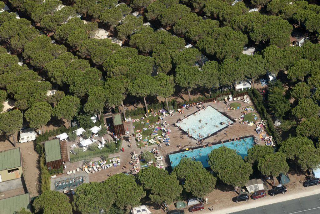 Oversigtsbillede af en campingplads med swimmingpool, omgivet af træer, campingvogne og telte.