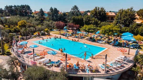 A swimming pool surrounded by sun loungers and umbrellas, with a view of trees and buildings in the background.