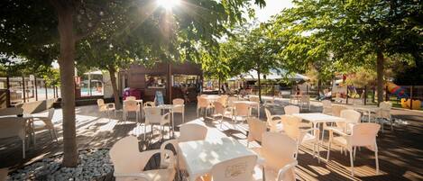 An outdoor seating area with tables and chairs, surrounded by trees and a wooden deck.
