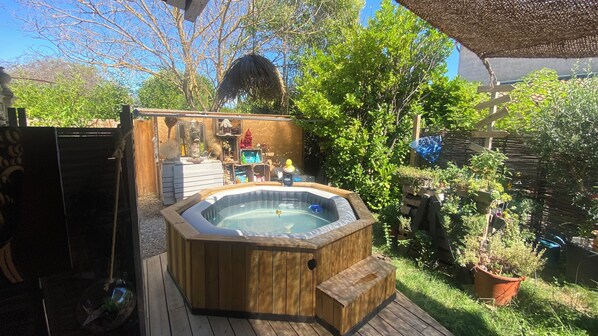 A wooden hot tub on a deck with a garden and various items in the background.