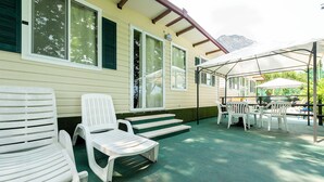 A covered patio with white plastic chairs and a table, a set of steps leading to a door, and a mountain in the background.