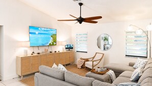 A living room with a television, a ceiling fan, a wooden cabinet, a round mirror, and a window with blinds. - Beautiful Home with Sauna & Heated Pool-Spa, 5min from Beaches, close to Airport (Lantana)