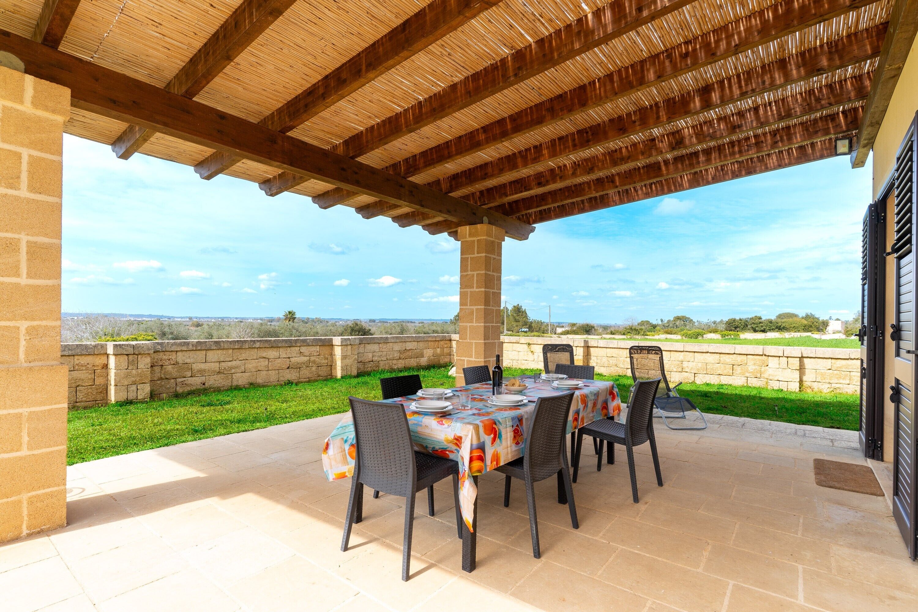 An outdoor patio with a table set for dining, surrounded by chairs, under a wooden pergola with a stone wall backdrop.