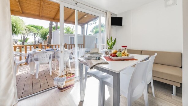 Un comedor luminoso y moderno con terraza de madera, mobiliario blanco y televisor montado en la pared.