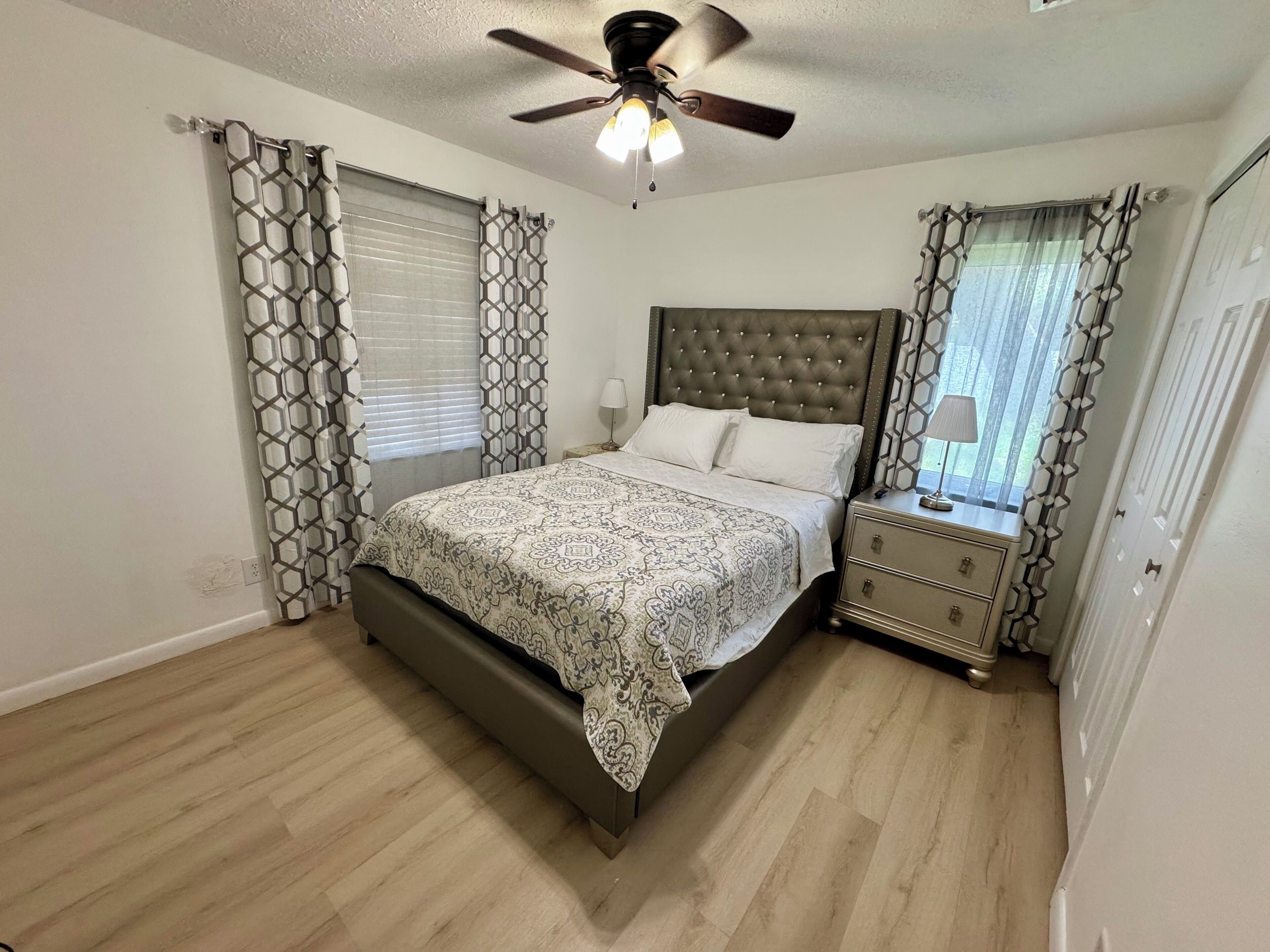 Een slaapkamer met een bed, nachtkastje, plafondventilator en decoratieve gordijnen.