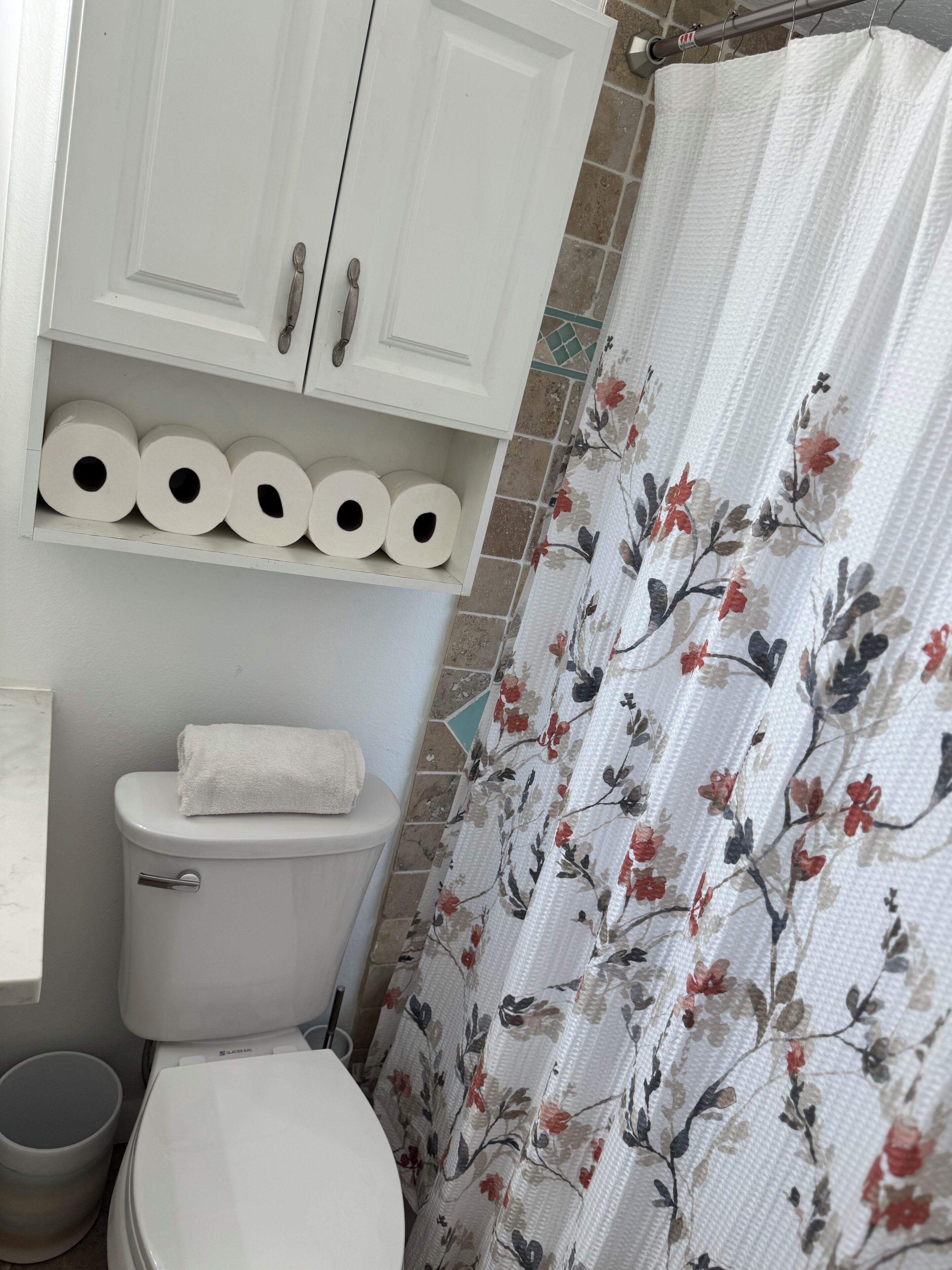 A bathroom with a toilet, a shelf with toilet paper, a shower curtain with a floral pattern, and a tiled shower wall.