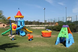 A playground with various play structures, including slides and climbing areas, surrounded by a fence and greenery.