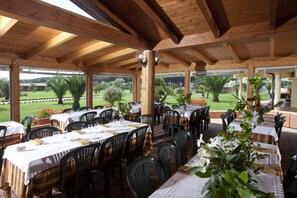 Un área de comedor al aire libre con mesas y sillas, vigas de madera y vistas a un jardín ajardinado.