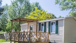 A mobile home with a covered porch and a small deck, surrounded by trees and greenery. - Ferienanlage Manacore Mobilehome Happy Easy für 6 Personen (Manaccora)