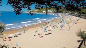 A beach with numerous umbrellas and sun loungers, a clear blue sea, and buildings in the background.