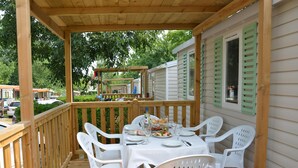 A covered outdoor dining area with a wooden deck, white plastic chairs, and a table set for a meal.