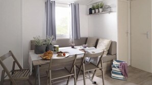 A dining area with a table, chairs, a bench, and a window with curtains. There are potted plants on a shelf above the bench.