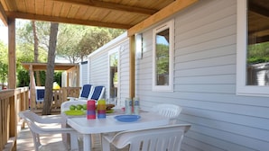 Una terraza techada al aire libre con una mesa blanca y sillas de plástico, barandal de madera y una casa móvil blanca al fondo.
