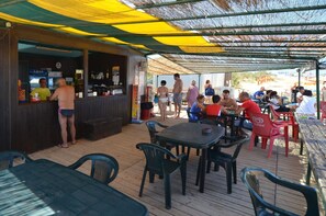 An outdoor bar area with wooden tables and chairs, a counter with a person serving, and a covered seating area with people dining.
