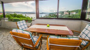 Una mesa de madera con mantel a cuadros, rodeada de sillas de madera con cojines a cuadros, en un balcón con vistas a un lago y colinas.
