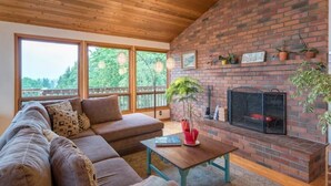 A cozy living room with a brick fireplace, wooden ceiling, and large windows. - Tabor Hillside Retreat over the trees, walk to trails, and Montavilla cafes.  (Portland)