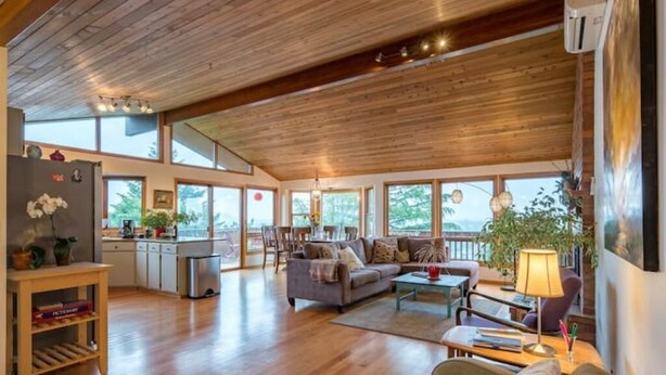 A spacious living area with wooden flooring, a sofa, a coffee table, and a dining table. There is a kitchen area with a refrigerator and a sink. Large windows provide natural light and views of the outdoors. - Tabor Hillside Retreat over the trees, walk to trails, and Montavilla cafes.  (Portland)