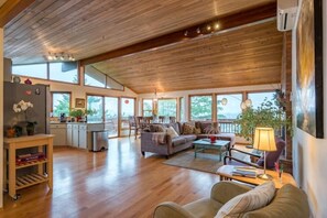 A spacious living area with wooden flooring, a sofa, a coffee table, and a dining table. There is a kitchen area with a refrigerator and a sink. Large windows provide natural light and views of the outdoors. - Tabor Hillside Retreat over the trees, walk to trails, and Montavilla cafes. (Portland)