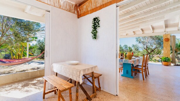 A room with a wooden table and bench, a hammock outside, and a view of greenery.