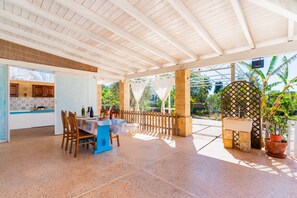 A covered patio with a table and chairs, a view of the garden, and a kitchen area.