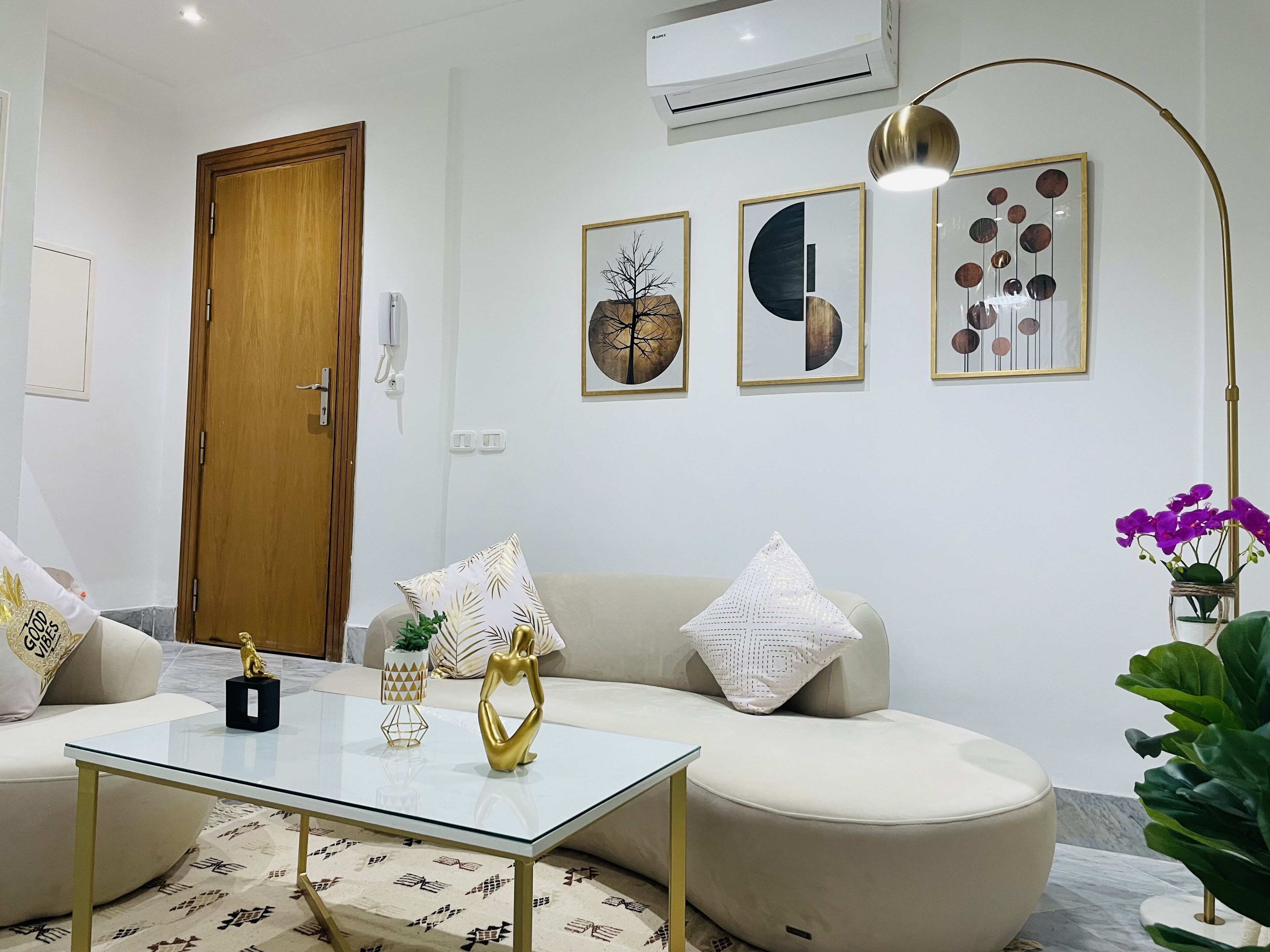 A modern living room with a glass coffee table, a beige sofa, and decorative pillows. There is a wooden door, an air conditioning unit, and wall art.
