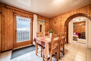 A wooden interior with a dining table, chairs, a potted plant, and a view through a door.